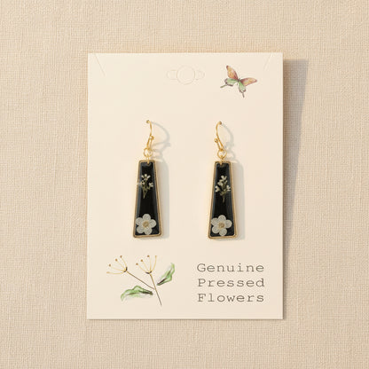 Black teardrop earrings with floral details on a beige card with botanical illustrations.