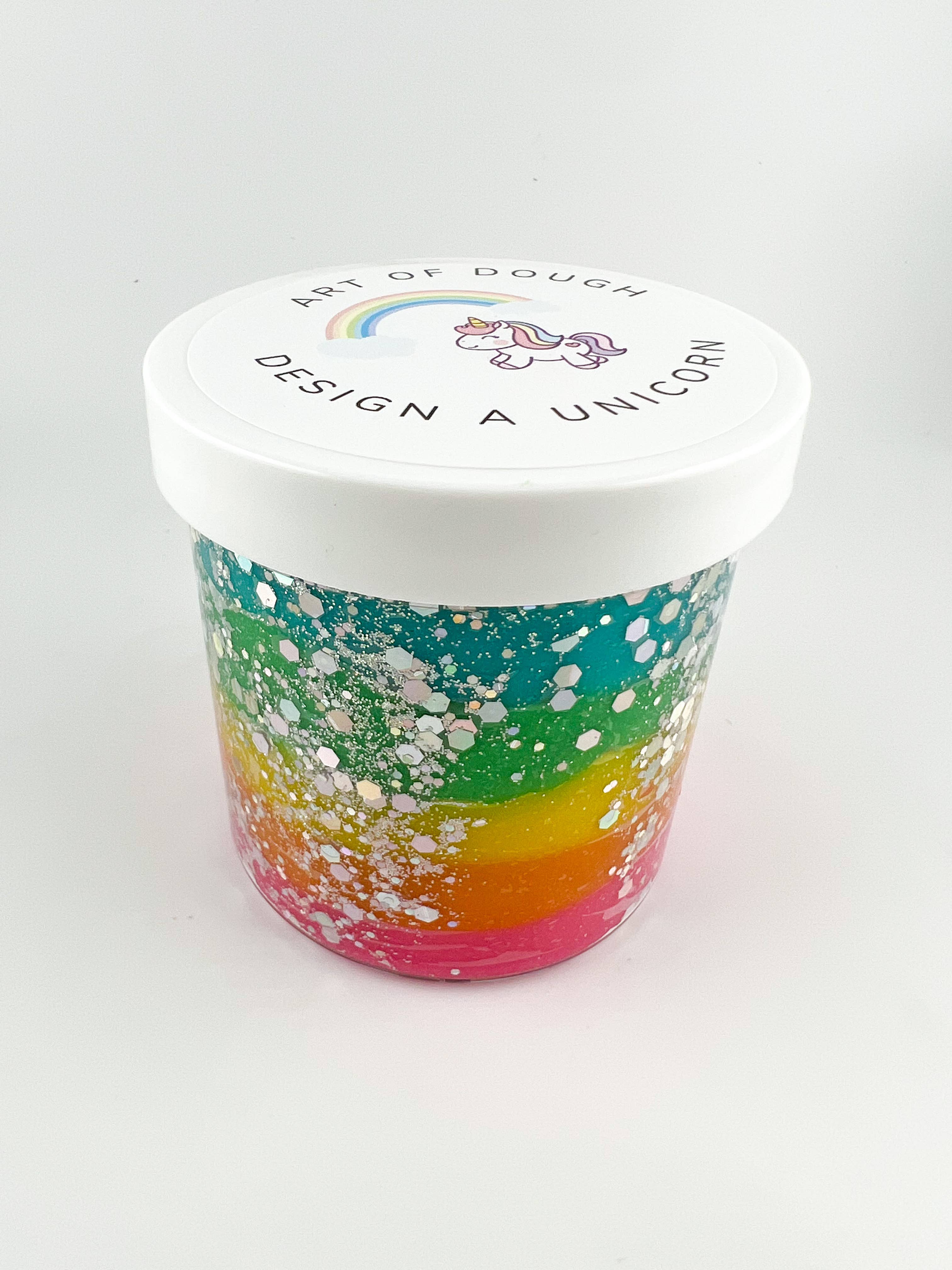 A 6oz jar containing glittery rainbow sensory dough, a unicorn figurine, a rainbow figurine, a unicorn cutter, and colorful gems.