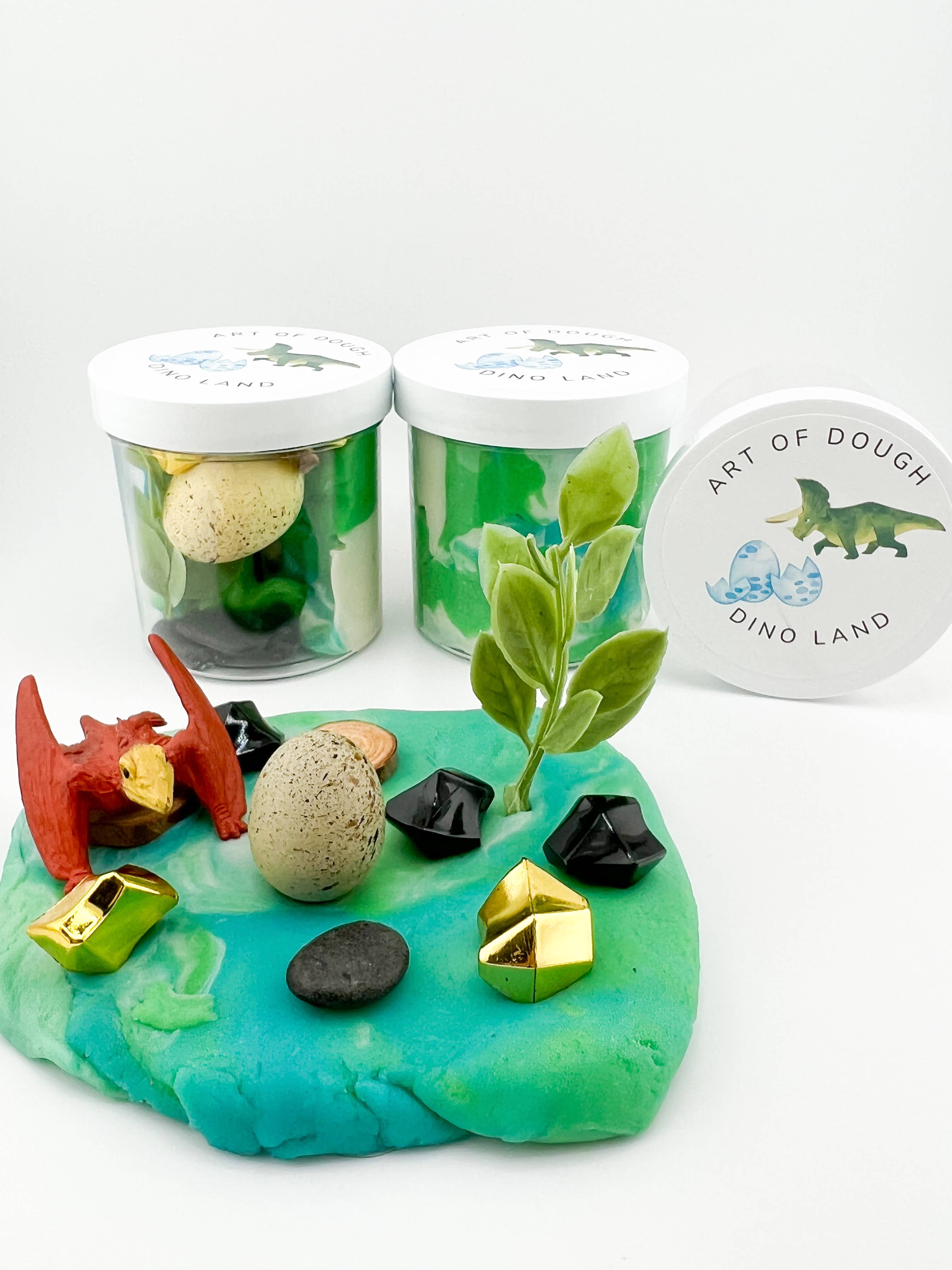 A 6oz jar featuring blue, white, and green swirled sensory dough, a dinosaur figurine, pebbles, a faux plant, and acrylic gems.