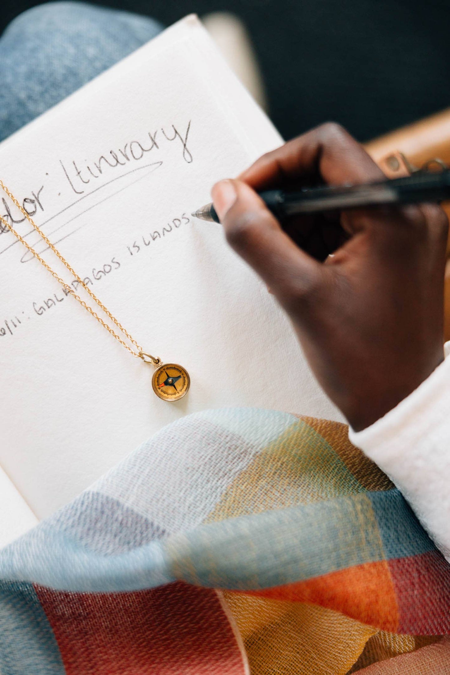 Person writing in a notebook with a pen, wearing a colorful garment and gold necklace.