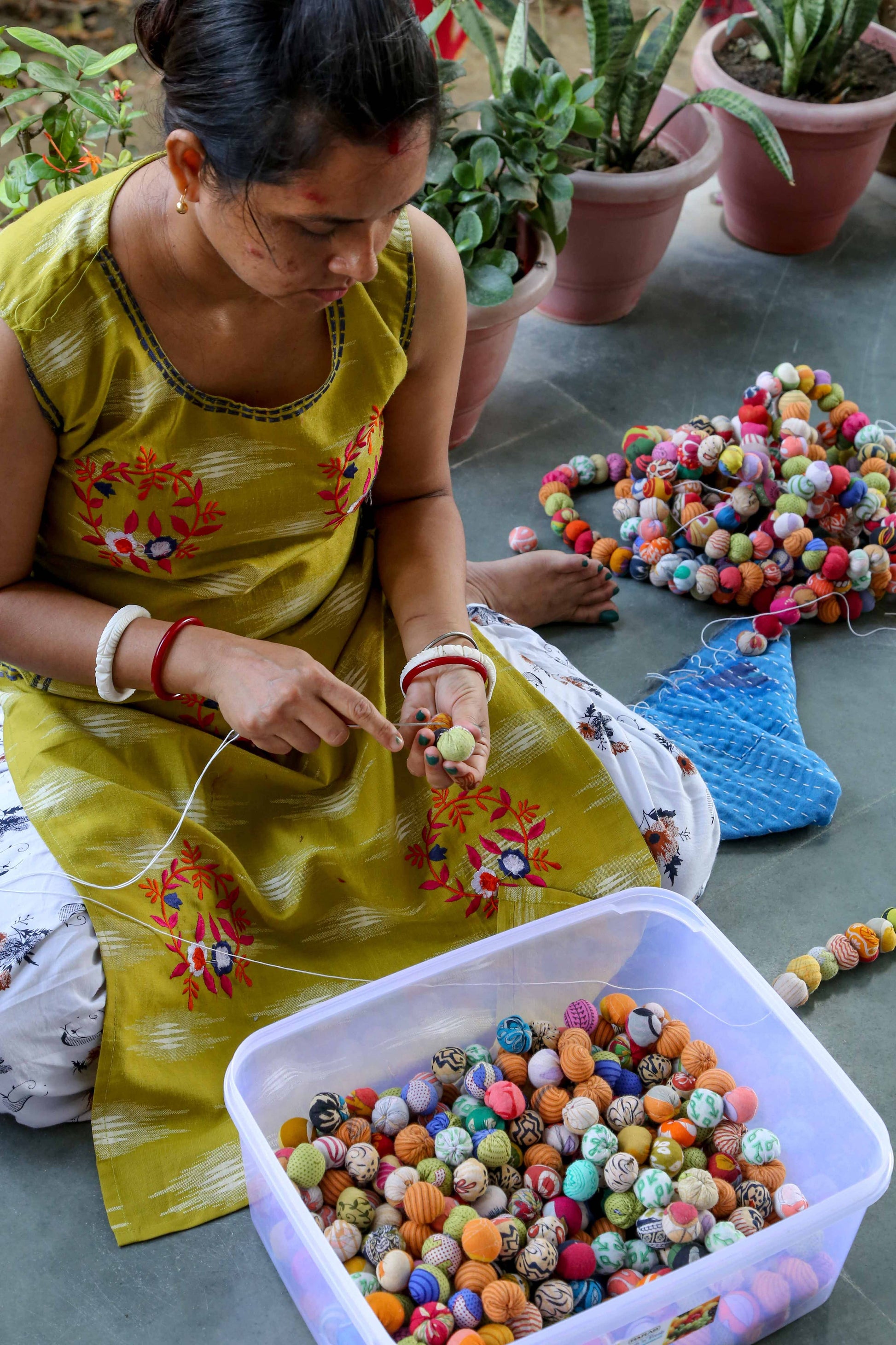 An Indian woman in a yellow dress sorting colorful beads with plants in the background.