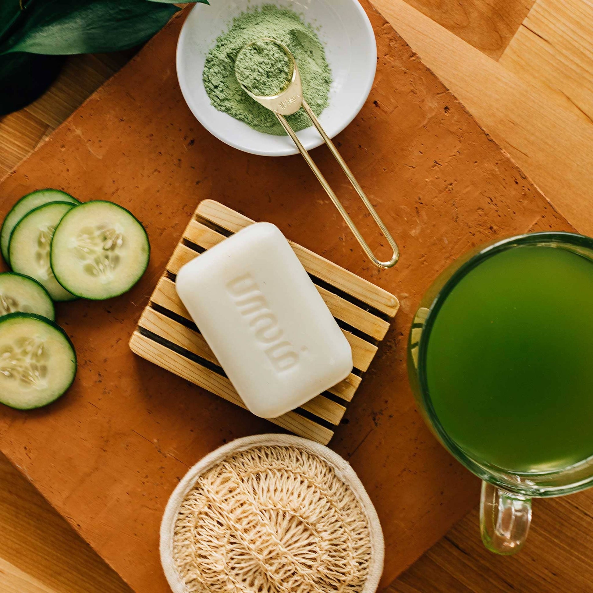 Bar of soap on a wooden coaster with cucumbers, a bowl of green powder, and a mug of green liquid on a wooden surface.