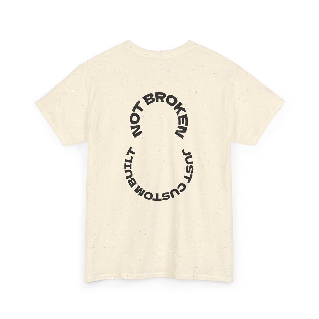 Beige t-shirt with black circular text that says, "Not Broken, Just Custom Built" design on a white background
