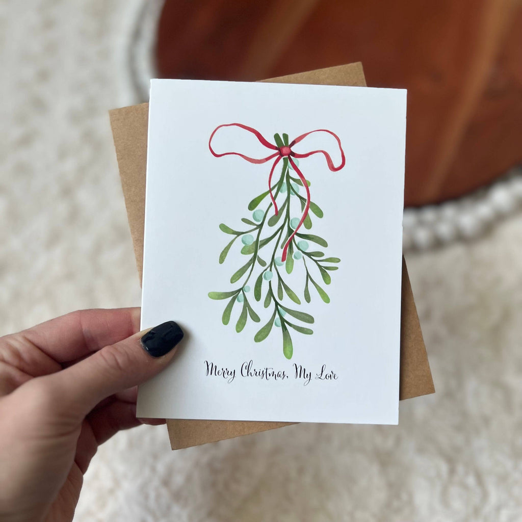 A hand holding a Christmas greeting card with an illustration of a mistletoe and the text 'Merry Christmas, My Love'. The card is paired with a rustic kraft envelope.