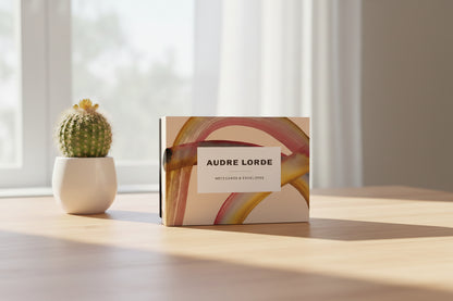 Packaging of Audre Lorde notecards and envelopes with abstract design on a desktop next to a plant with a window in the background