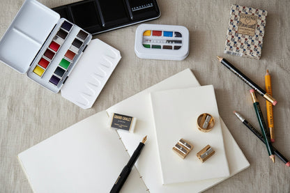 Art supplies including watercolor paints, pencils, and a notebook on a wooden surface.