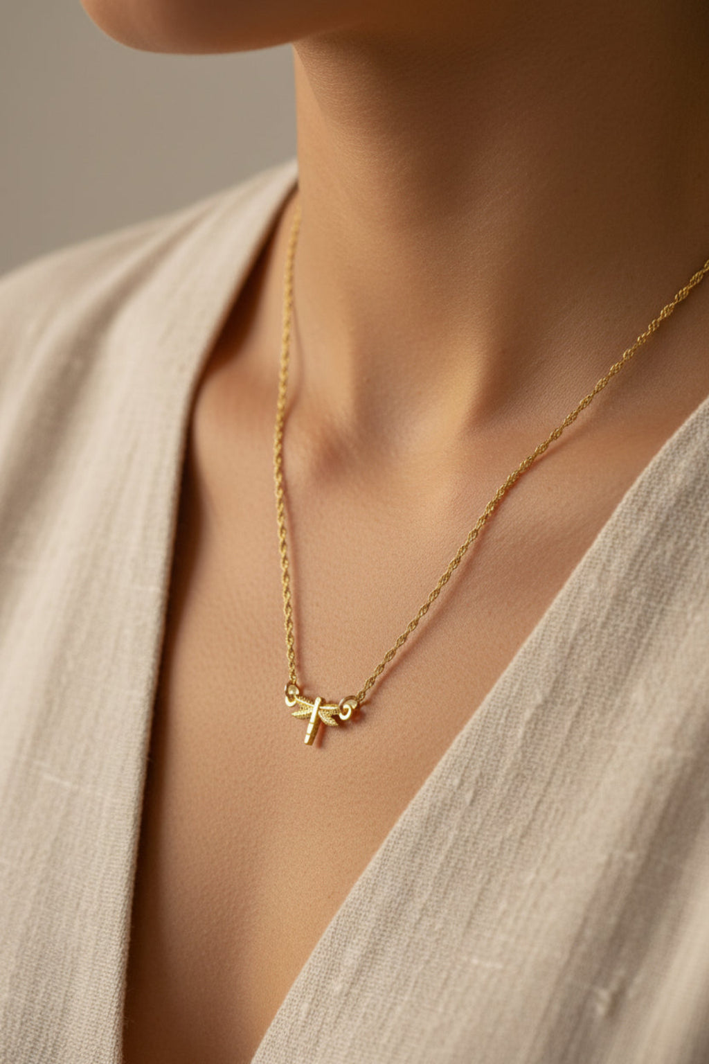 Gold necklace with a small dragonfly pendant being worn with natural garments and a light gray background