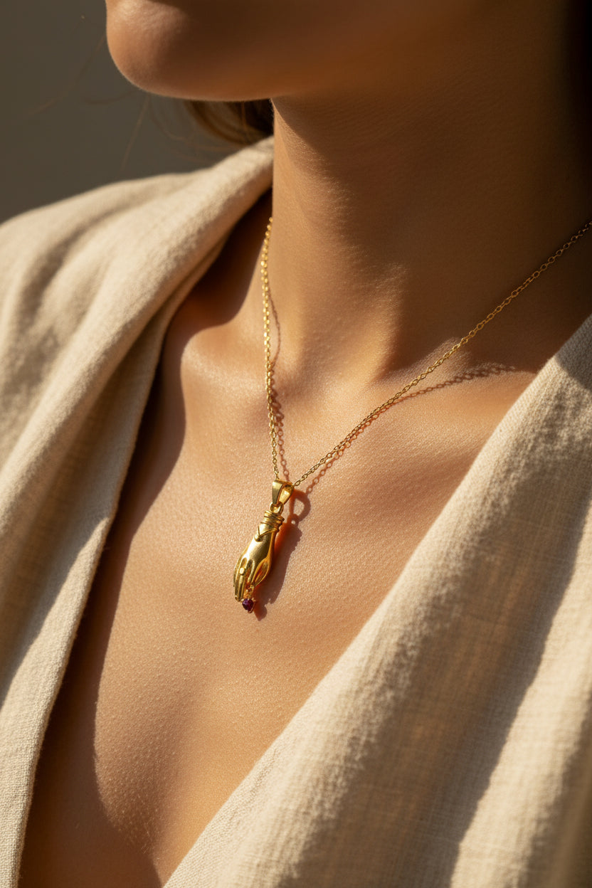 Gold pendant shaped like a hand holding a red gemstone on a gold chain being worn in a lifestyle shot