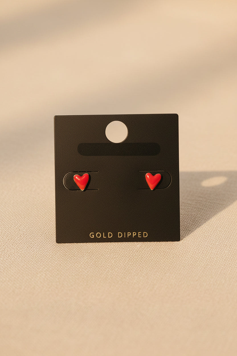 Red heart-shaped earrings on a black card with 'Gold Dipped' text, placed on a beige fabric background.