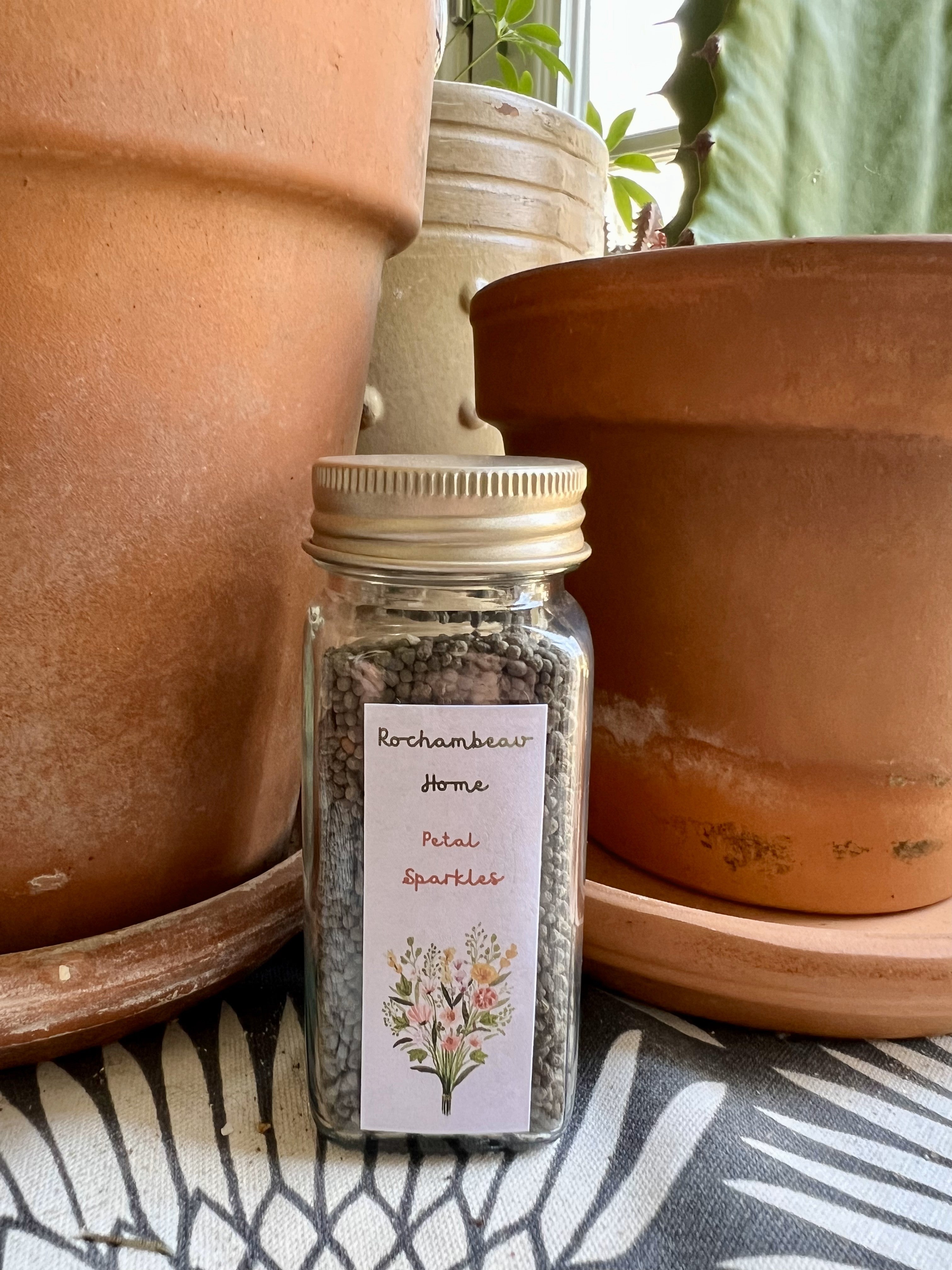 A reusable glass jar with a gold tin lid labeled "Petal Sparkles," filled with a colorful mix of wildflower seeds.
