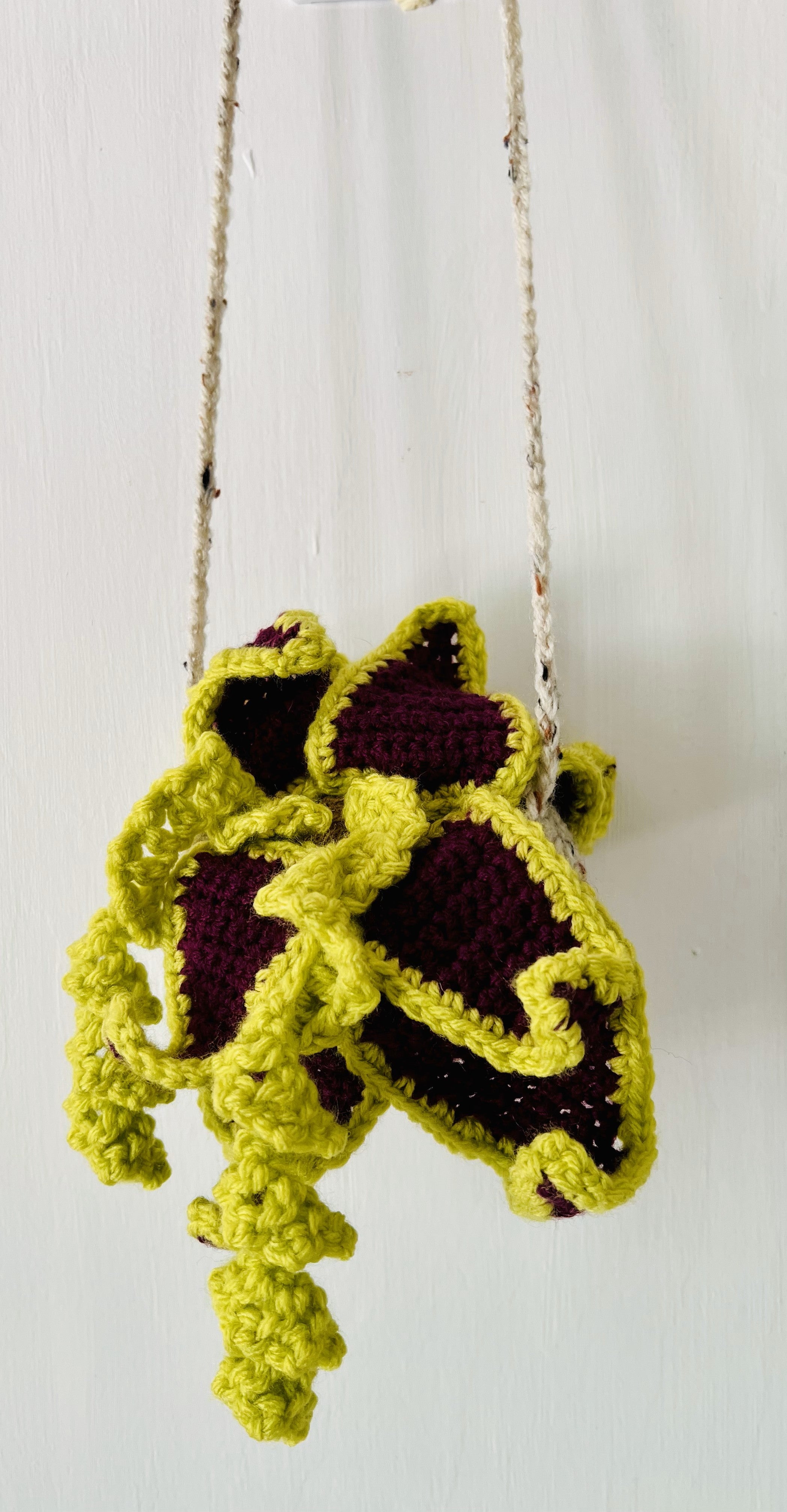 Oversized crocheted air plant with maroon and green yarn in a neutral pot on wooden shelf