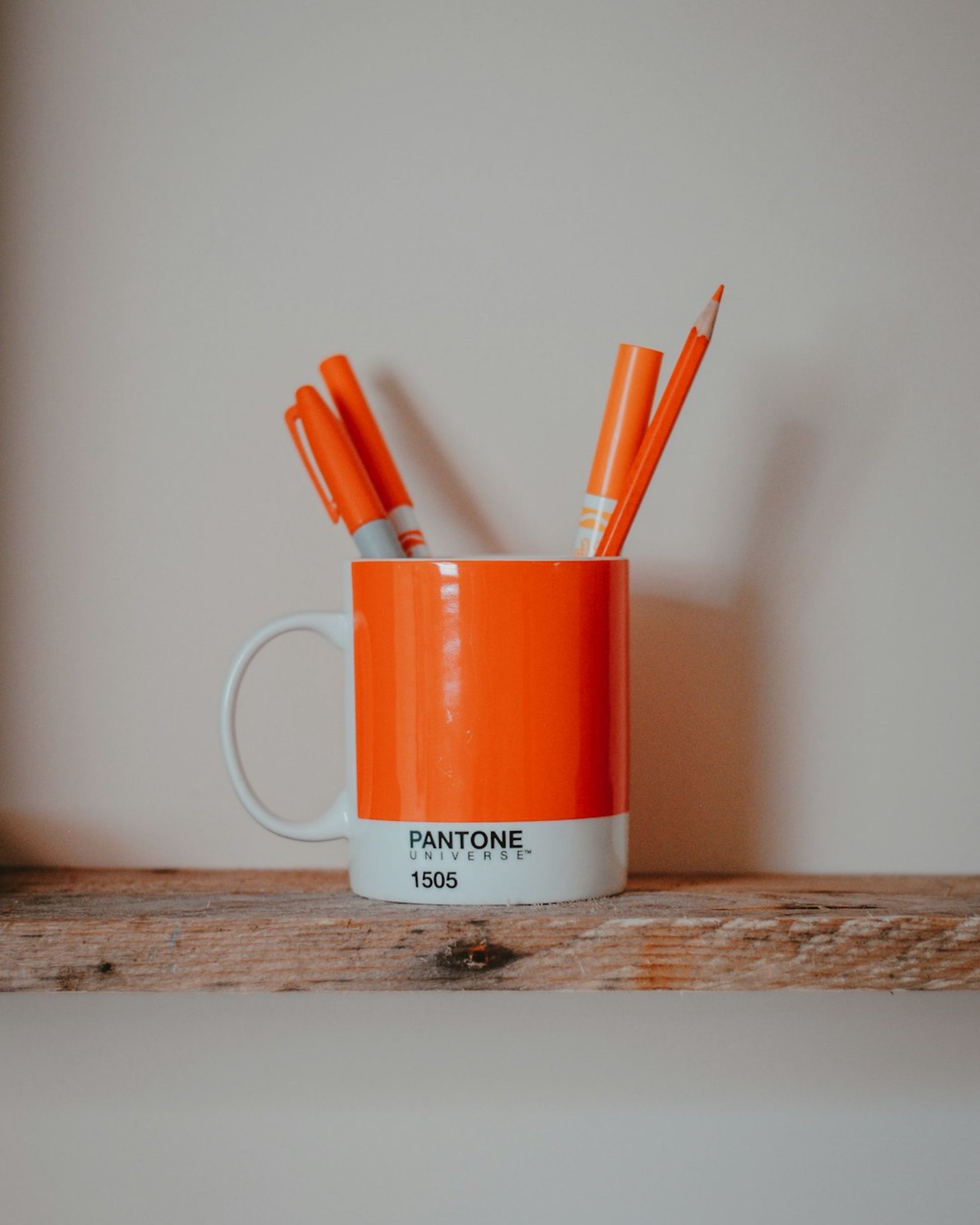 Orange mug with Pantone color swatch containing orange pens and pencils on a wooden surface.