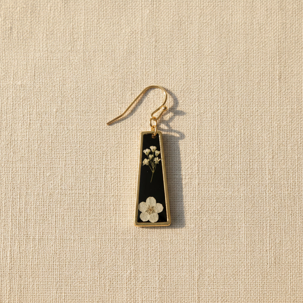 A single drop earring with a black background and small pressed white flowers pressed in resin