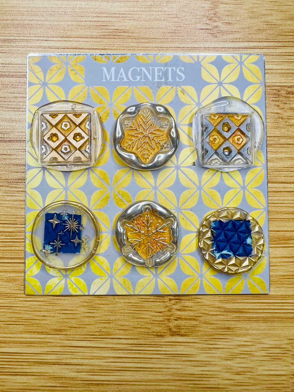 Set of six decorative magnets on a patterned card with 'Magnets' text, on a wooden surface.