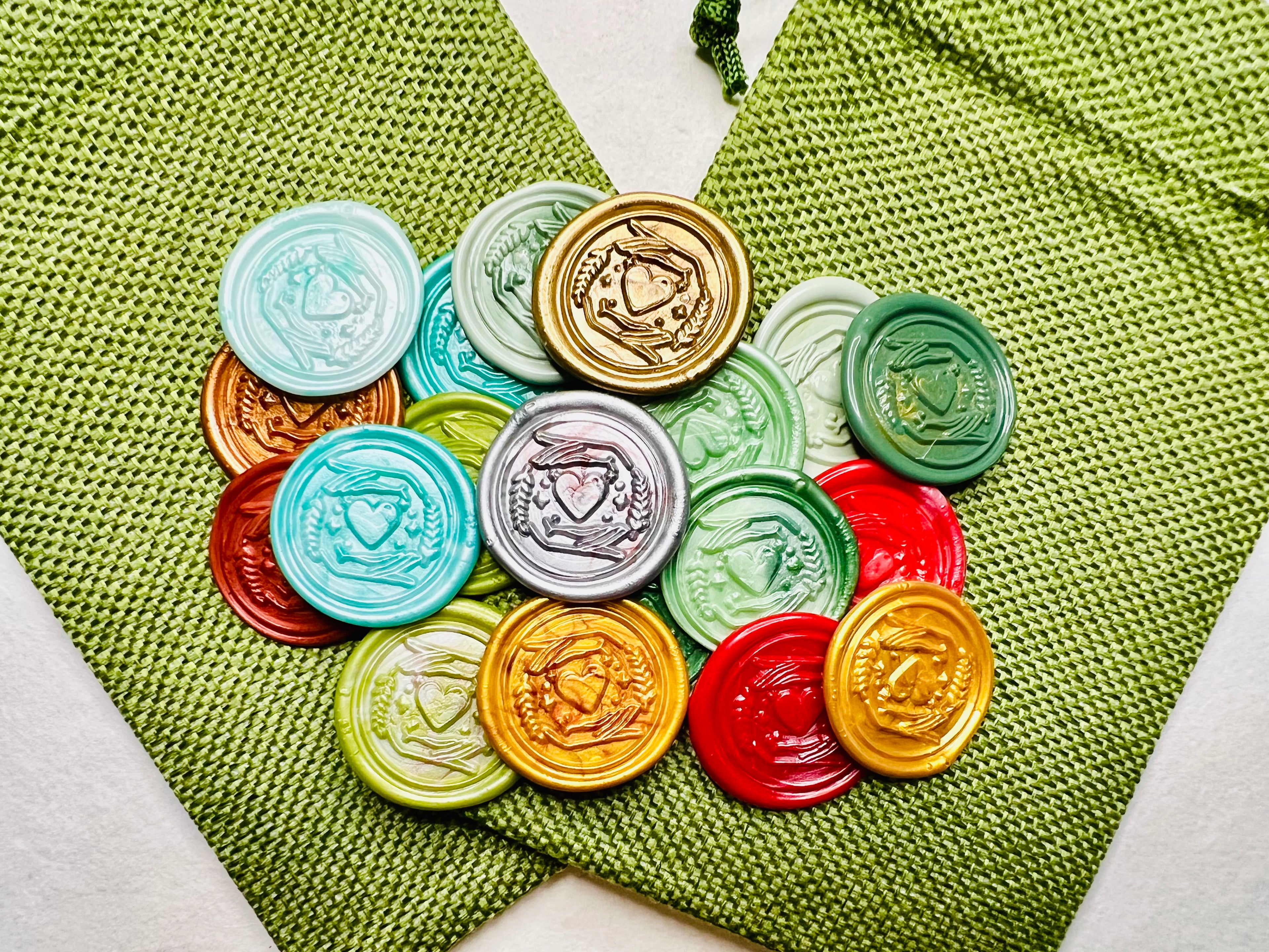 Holiday Heart in Hand wax seal stickers created by Rochambeau Home in various colors  being showcased on green burlap bags.