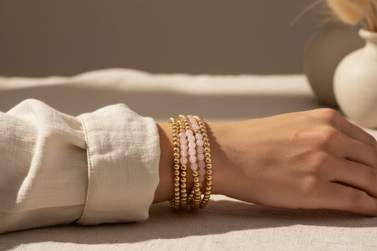 Stack of gold beaded bracelets shown here on a person's wrist with a beige top and neutral background