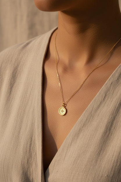 Gold necklace with a compass pendant being worn with neutral toned garments on a neutral background