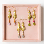 A set of golden brass hand charm earrings displayed on a pink tray, each earring featuring a trio of Czech glass beads in different colors.