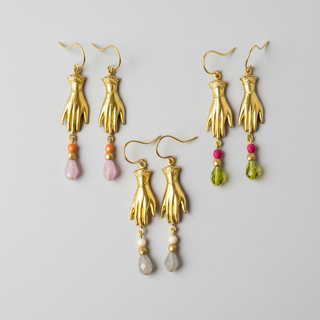 A set of golden brass hand charm earrings displayed on a pink tray, each earring featuring a trio of Czech glass beads in different colors.