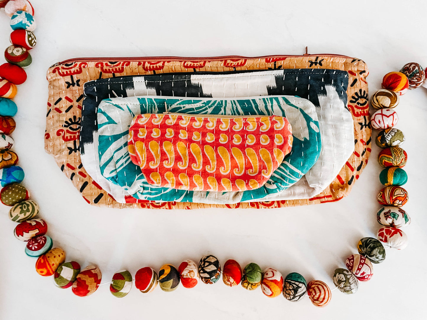 Stack of colorful pouches with patterned contents on a white background and sari garland