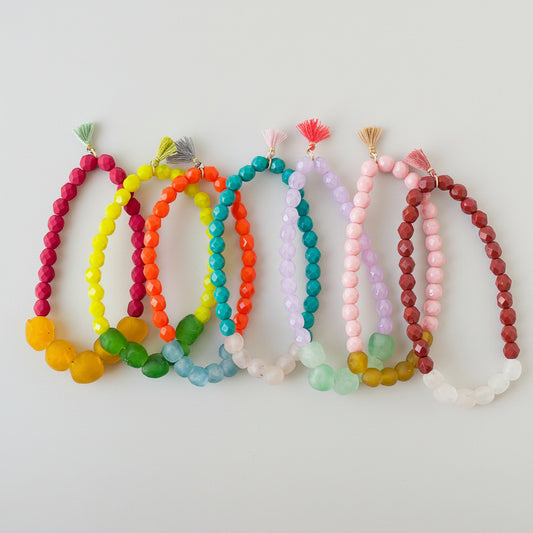 A collection of stretchy bracelets with recycled African glass beads in various bright colors, each featuring a tiny tassel.