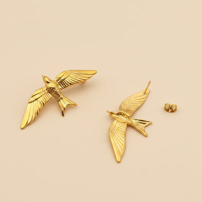 Gold bird-shaped earrings on a light surface with a blurred background of shelves and bags.