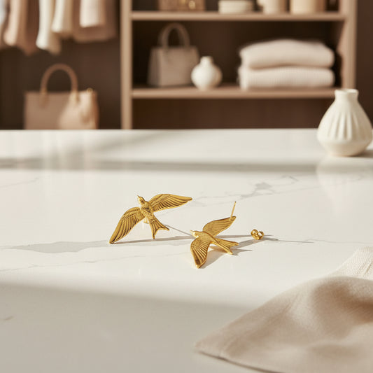 Gold bird-shaped earrings on a light surface with a blurred background of shelves and bags.