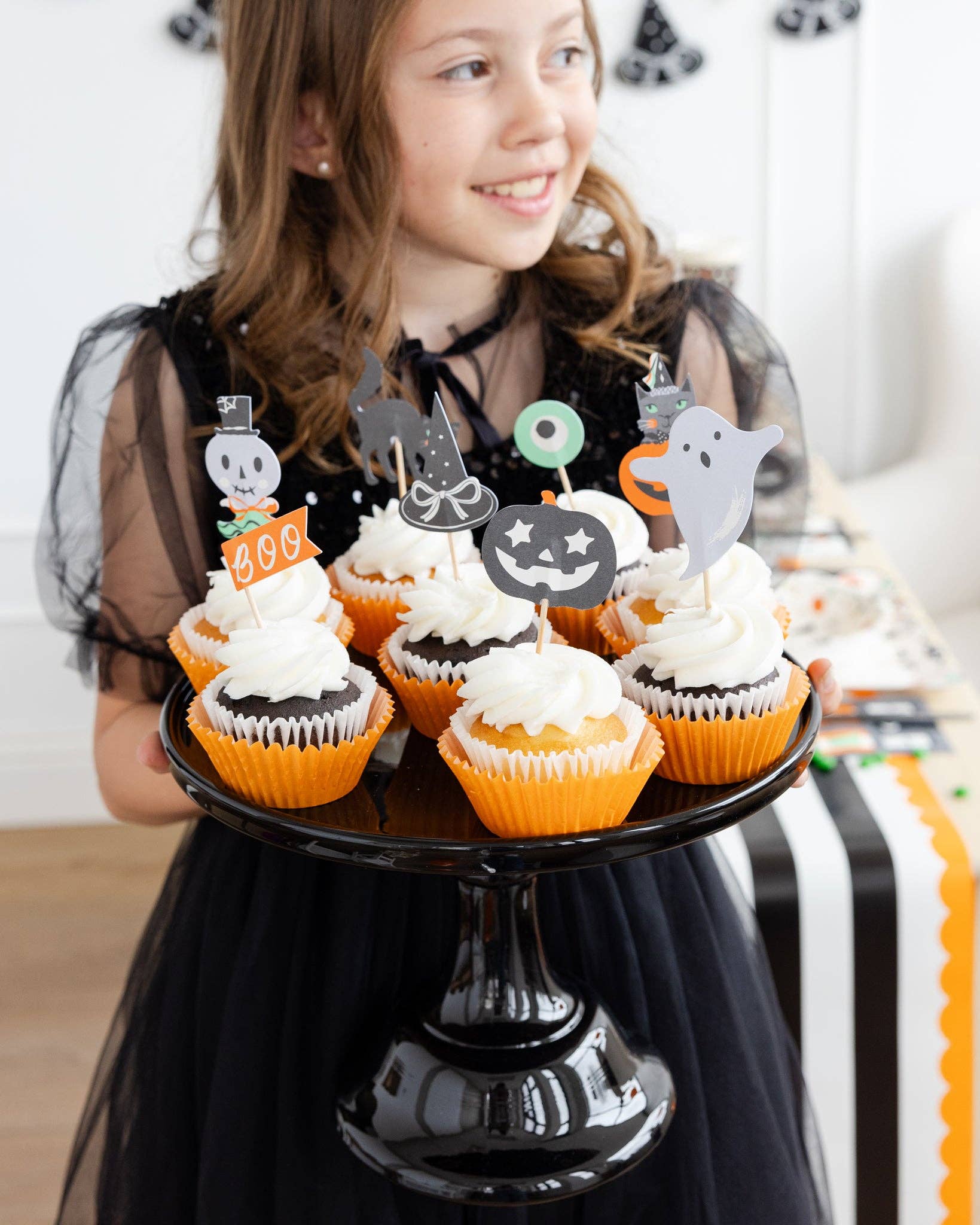 Girl holding a black cake stand with Halloween-themed cupcakes.
