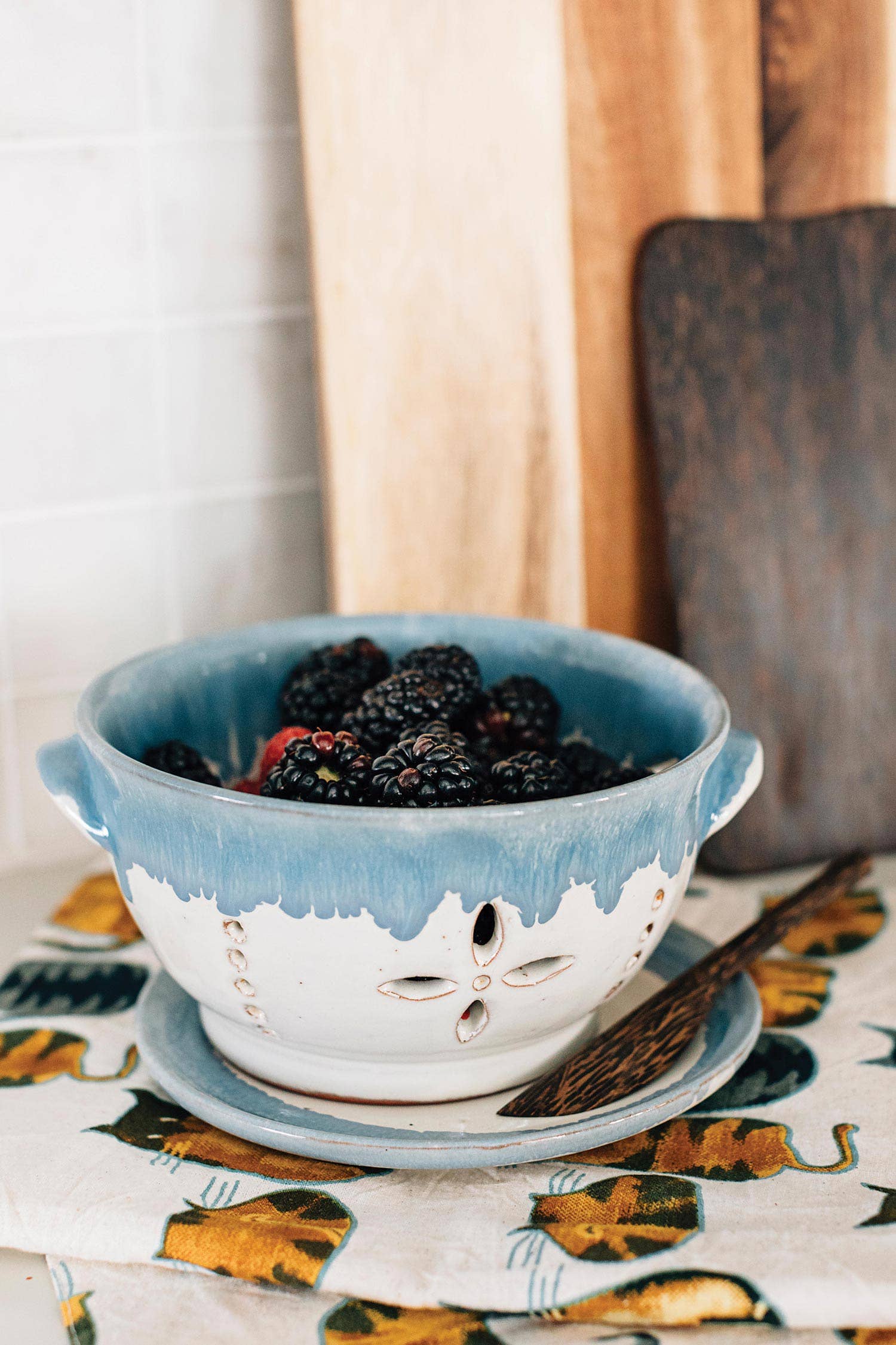 Blue ceramic colander filled with blackberries on a patterned tablecloth.