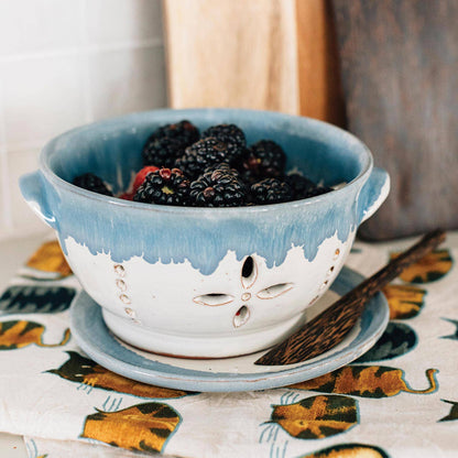 Blue ceramic colander filled with blackberries on a patterned tablecloth.