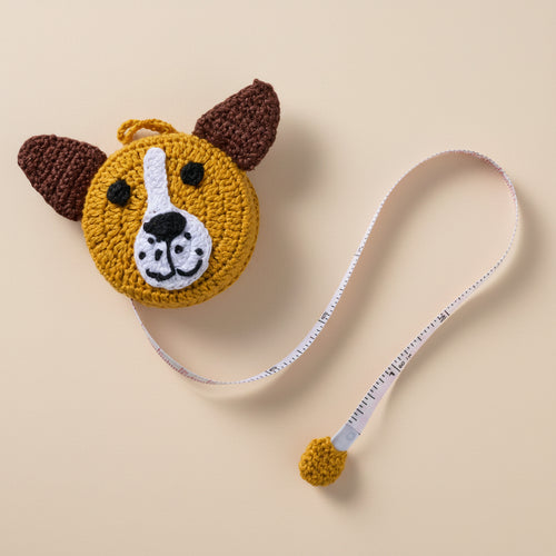 Crochet dog measuring tape with floppy 3D ears and smiling face, shown on a craft table