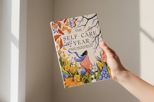 Hand holding a book titled 'The Self-Care Year' with a colorful cover against a neutral background