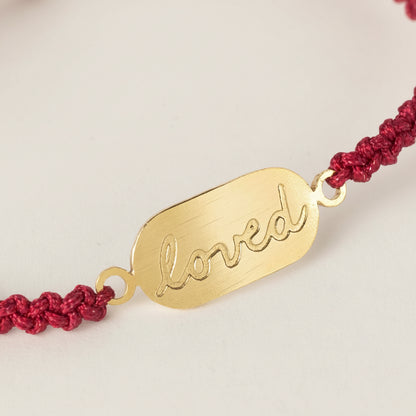 Close-up of red nylon cord bracelet featuring “You Are Loved” brass detail