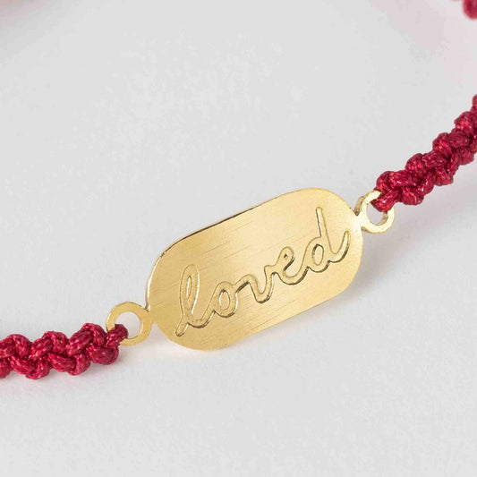 Close-up of red nylon cord bracelet featuring “You Are Loved” brass detail