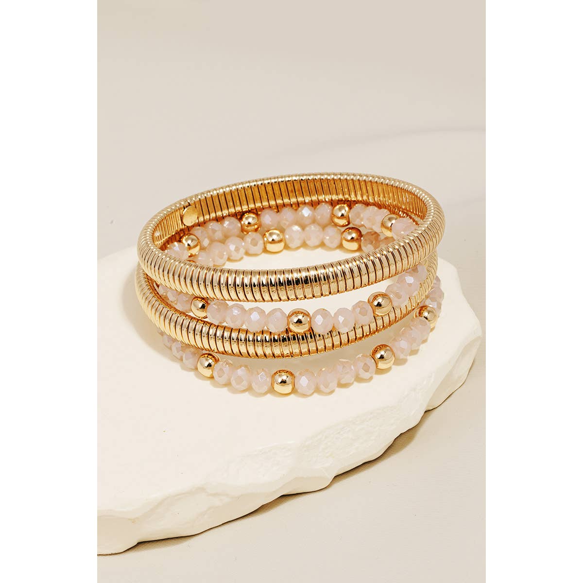 A stack of gold-colored metallic coil and beaded bracelets on a white cylindrical display stand.