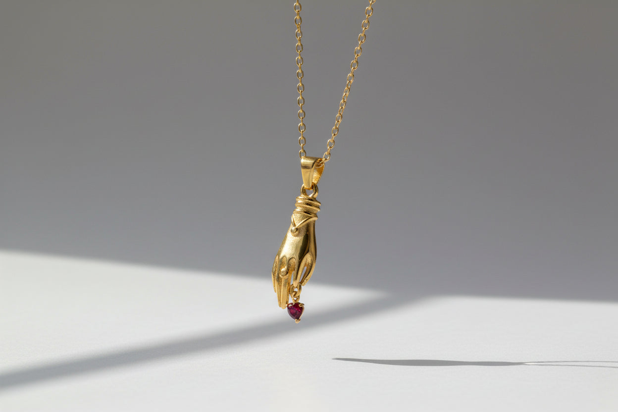 Gold necklace with a hand and heart pendant held in a hand against a gray background