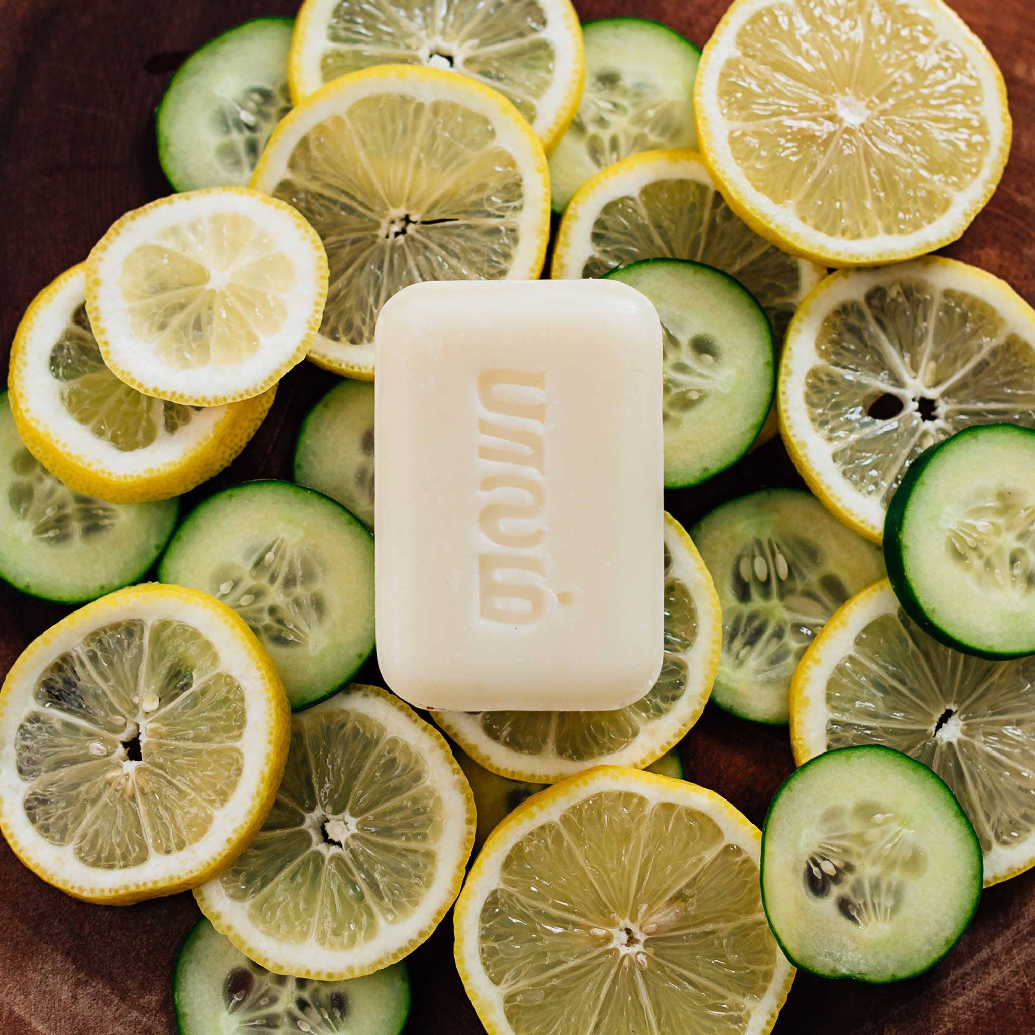 Bar of soap surrounded by sliced lemons and cucumbers on a wooden surface
