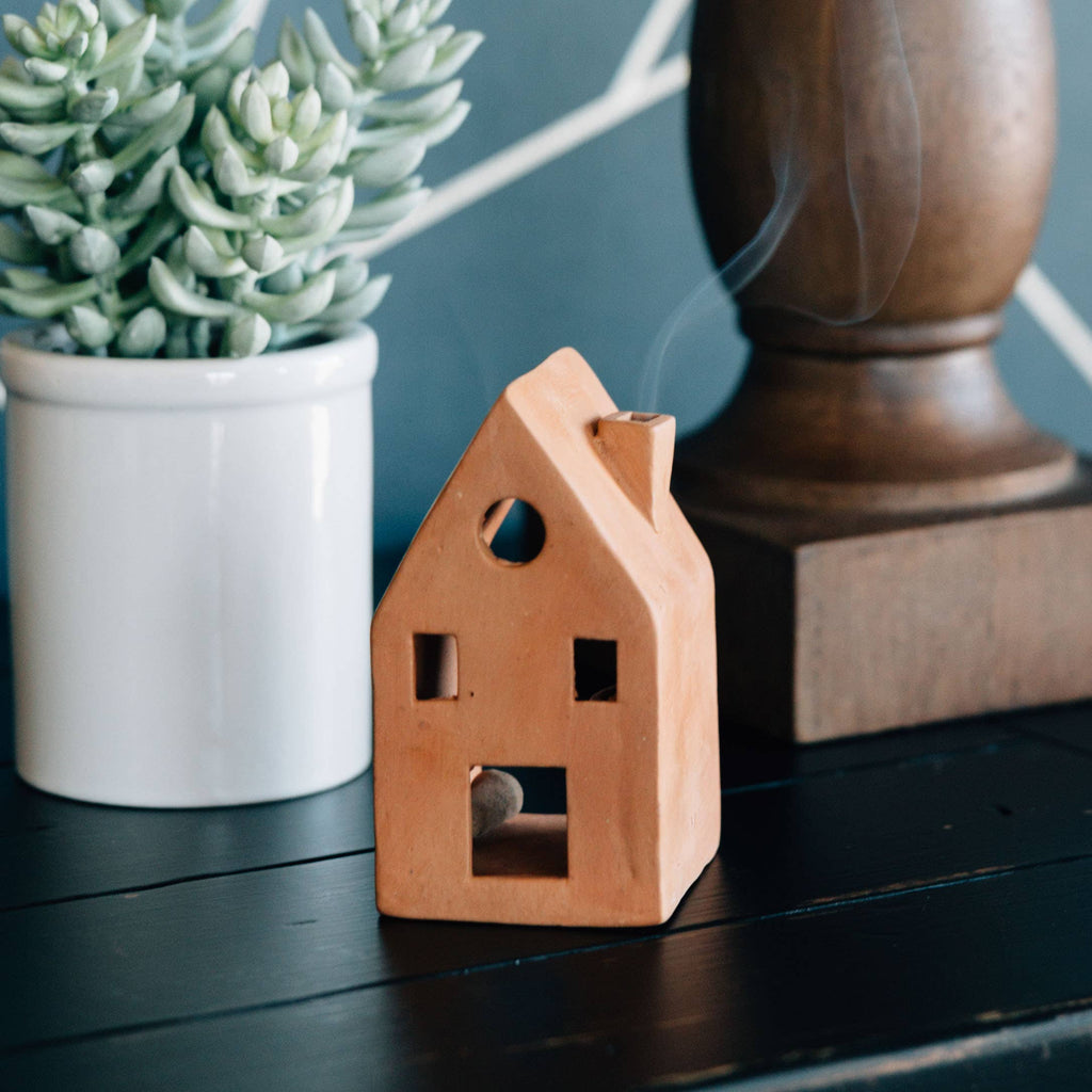 Terracotta house-shaped incense burner with smoke, next to a potted plant and wooden object on a dark surface.