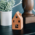 Terracotta house-shaped incense burner with smoke, next to a potted plant and wooden object on a dark surface.